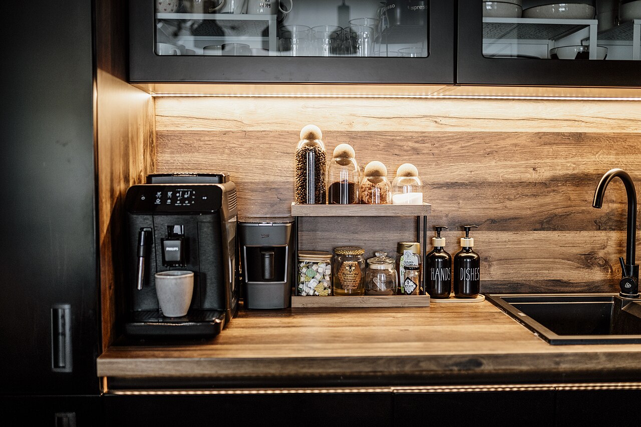 A home coffee station on a wooden counter: a black bean-to-cup coffee machine with a cup under the spout, a smaller grey appliance beside it, and jars of coffee beans on a shelf above